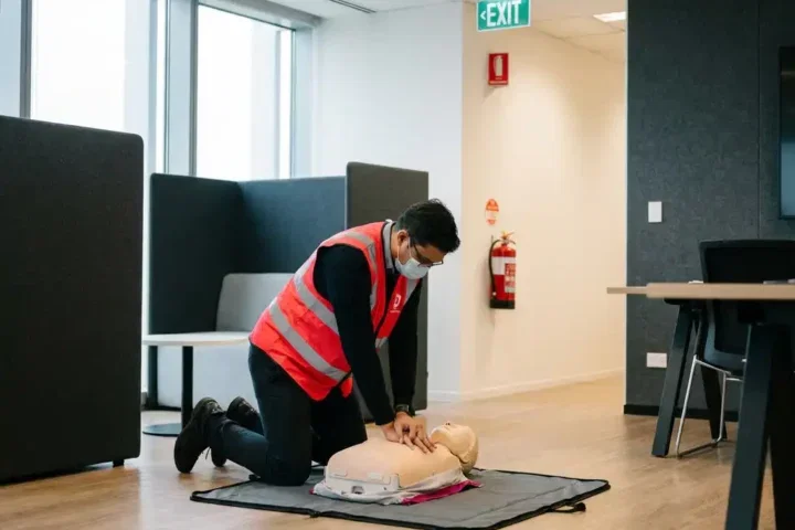 BHV-medewerker in rood veiligheidsvest oefent hartmassage op reanimatiepop in moderne kantoortrainingsruimte.