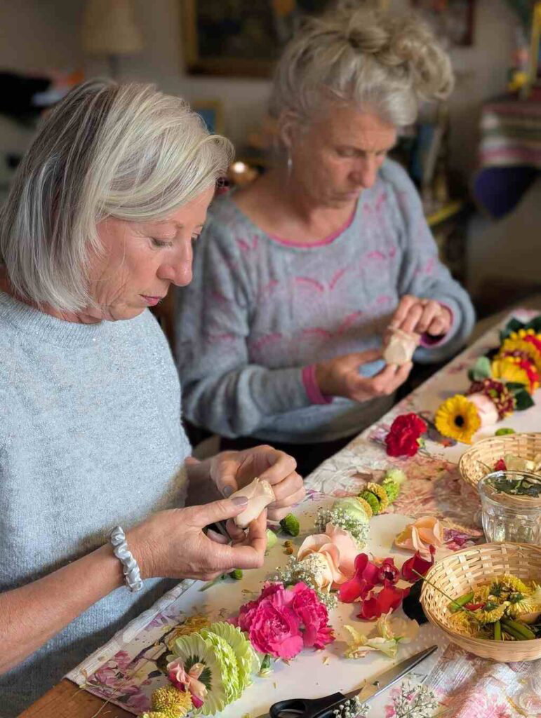 Shakti Bloemenmala Tonny Bol twee cursisten vrouwen gefocust bezig kleurige bloemen rijgen bloemenslingers creëren tafel bloemblaadjes