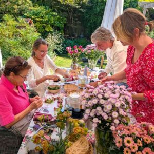 Shakti Bloemenmala Tonny Bol Shakti Flower Meeting in bloementuin aan tafel bloemenslingers maken verse bloemen vrolijke groep vrouwen