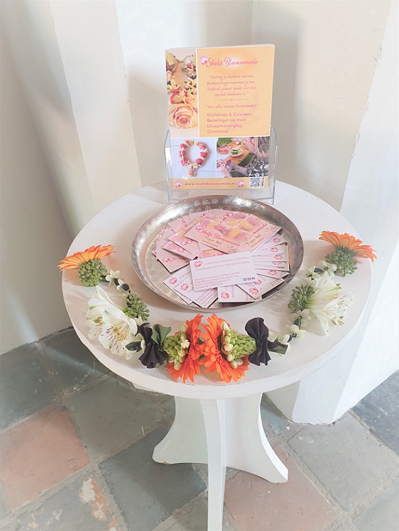 Shakti Bloemenmala, Tonny Bol, bloemenslinger met oranje en witte bloemen op tafel met visitekaartjes en flyers